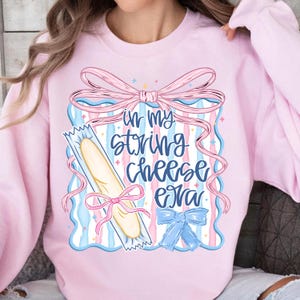 May include: Pale pink sweatshirt with a graphic design. The design includes a string cheese package, bows, and the text "in my string cheese era" in blue script. The design includes pink, blue, and white colors.