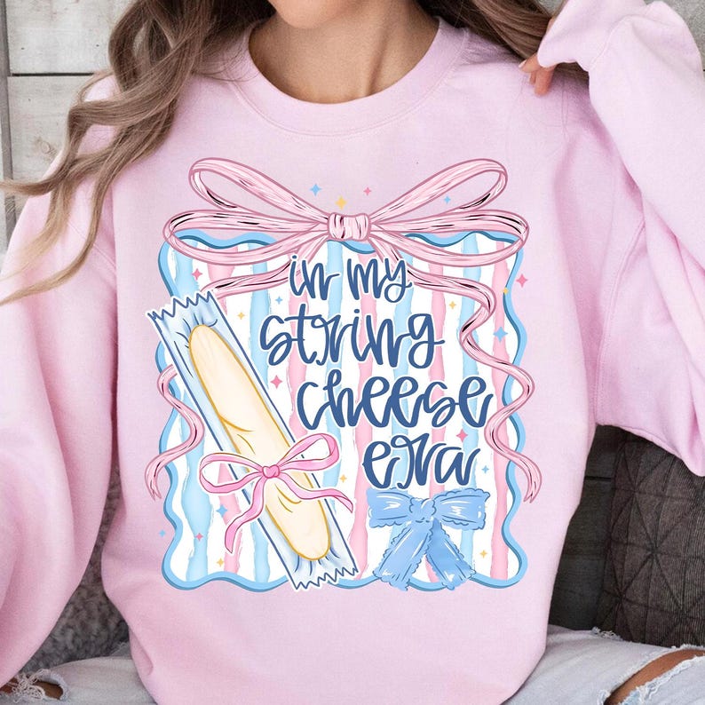 May include: Pale pink sweatshirt with a graphic design. The design includes a string cheese package, bows, and the text "in my string cheese era" in blue script. The design includes pink, blue, and white colors.