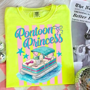 May include: A neon yellow t-shirt with a graphic that says "Pontoon Princess". The graphic features a cartoon pontoon boat with a teal canopy, pink and teal accents, and blue stars. The shirt has a Comfort Colors tag.