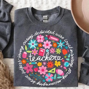 May include: A dark gray sweatshirt with a circle design featuring colorful flowers, school supplies, and the word "teacher" surrounded by a phrase that reads "smart - strong - brave - loving - caring - dedicated - wise - loyal - inspiring - fun".