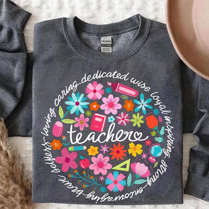 May include: A dark gray sweatshirt with a circle design featuring colorful flowers, school supplies, and the word "teacher" surrounded by a phrase that reads "smart - strong - brave - loving - caring - dedicated - wise - loyal - inspiring - fun".