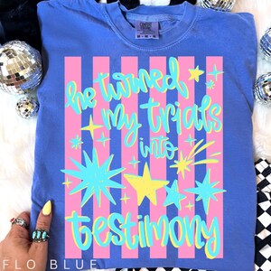 May include: Flo Blue Comfort Colors t-shirt with the text "He turned My trials into testimony" in a colorful design. The design includes pink stripes, stars, and script lettering.