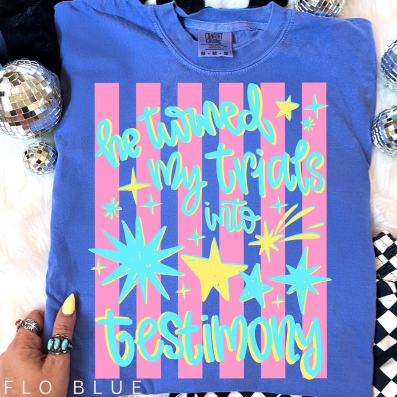 May include: Flo Blue Comfort Colors t-shirt with the text "He turned My trials into testimony" in a colorful design. The design includes pink stripes, stars, and script lettering.