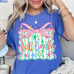 May include: Blue t-shirt with a colorful graphic. The graphic features a pink bow, green and pink stars, and the word "Hallelujah" in a multi-colored, whimsical font. The shirt is a solid blue color.