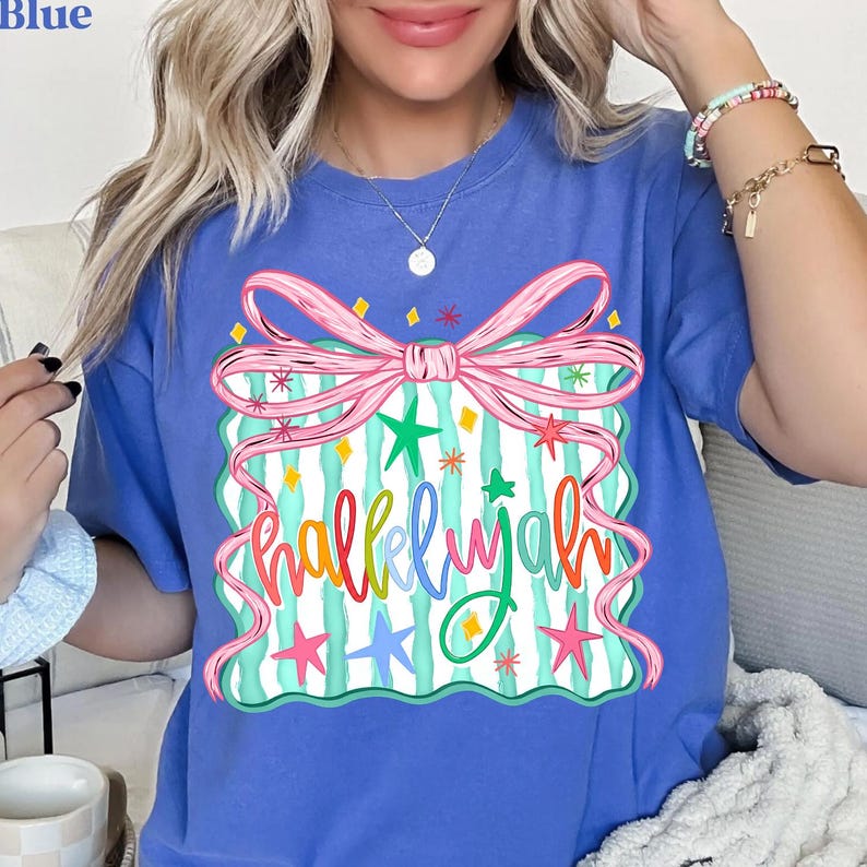 May include: Blue t-shirt with a colorful graphic. The graphic features a pink bow, green and pink stars, and the word "Hallelujah" in a multi-colored, whimsical font. The shirt is a solid blue color.