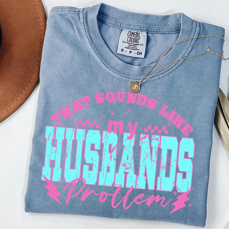 May include: A blue Comfort Colors t-shirt with the text "THAT SOUNDS LIKE MY HUSBANDS Problem" in pink and turquoise. A gold necklace with a pendant is draped across the shirt.
