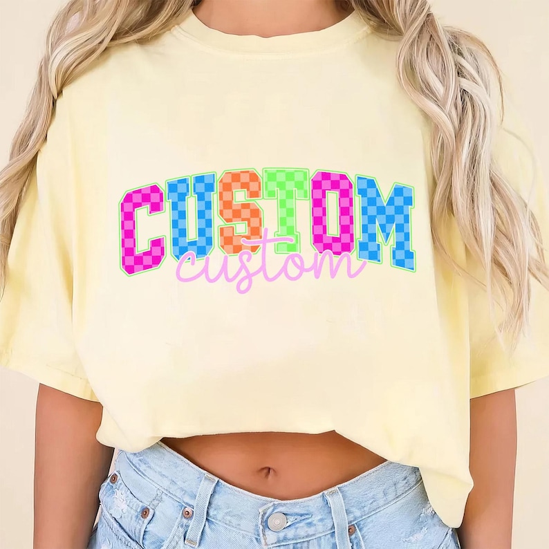 May include: A light yellow t-shirt with the word "CUSTOM" in a colorful, checkered design. The word "custom" is written in a cursive font below the main text. The shirt is made of a soft material.