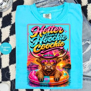 May include: Lagoon blue t-shirt with a graphic of a Highland cow wearing sunglasses and a pink cowboy hat, floating in a colorful inner tube. The text "Hooter than a Hoochie Coochie" is displayed above the cow.