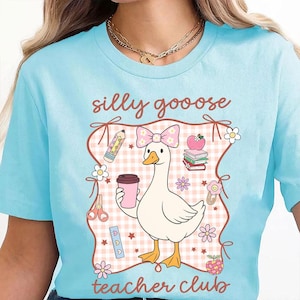 May include: Light blue t-shirt with a whimsical graphic featuring a goose wearing a bow and holding a coffee cup. The design includes the words "silly goooose teacher club" and school-themed illustrations like pencils, books, and flowers.