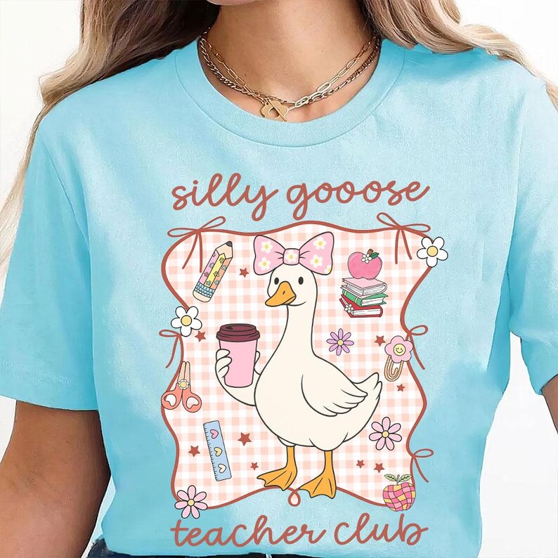 May include: Light blue t-shirt with a whimsical graphic featuring a goose wearing a bow and holding a coffee cup. The design includes the words "silly goooose teacher club" and school-themed illustrations like pencils, books, and flowers.