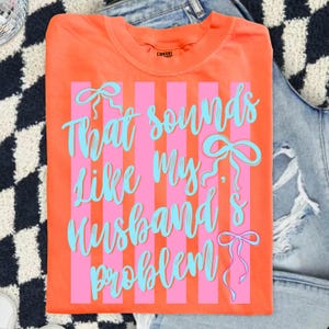 May include: Orange t-shirt with pink vertical stripes and the text "That sounds like my husband's problem" in turquoise script. The shirt has a crew neck and is folded. The shirt is on a denim background.