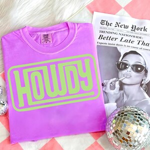 May include: A lilac-colored t-shirt with the word "HOWDY" in a lime green, outlined font. The shirt is a Comfort Colors brand. A black and white newspaper and a disco ball are in the background.