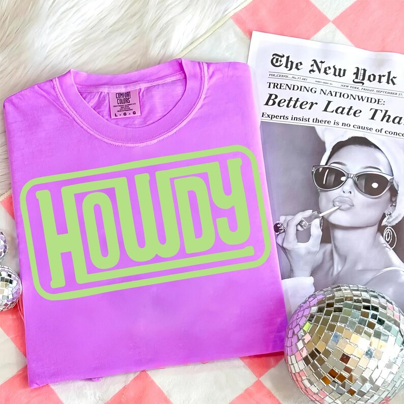 May include: A lilac-colored t-shirt with the word "HOWDY" in a lime green, outlined font. The shirt is a Comfort Colors brand. A black and white newspaper and a disco ball are in the background.