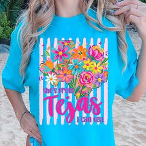 May include: A turquoise t-shirt featuring a floral Texas outline design with the text "She's from Texas I can tell." The design includes pink, orange, yellow, and blue flowers, and pink vertical stripes.