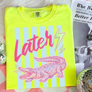 Pode incluir: Camiseta Comfort Colors amarelo neon com um gr&aacute;fico rosa e azul. O design apresenta a palavra "Later" em rosa, um jacar&eacute; rosa e azul e raios verdes. A camisa &eacute; tamanho L-G.