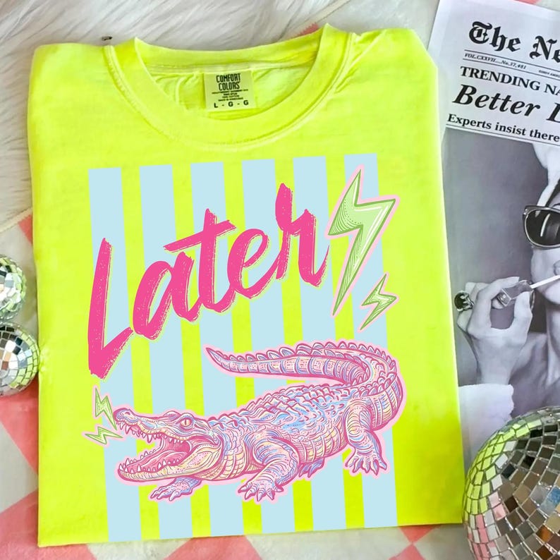 Pode incluir: Camiseta Comfort Colors amarelo neon com um gr&aacute;fico rosa e azul. O design apresenta a palavra "Later" em rosa, um jacar&eacute; rosa e azul e raios verdes. A camisa &eacute; tamanho L-G.