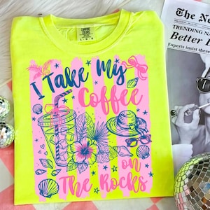 May include: Neon yellow t-shirt with a graphic design that says "I Take My Coffee on The Rocks." The design includes a coffee cup, hat, sunglasses, and tropical flowers in pink and blue.
