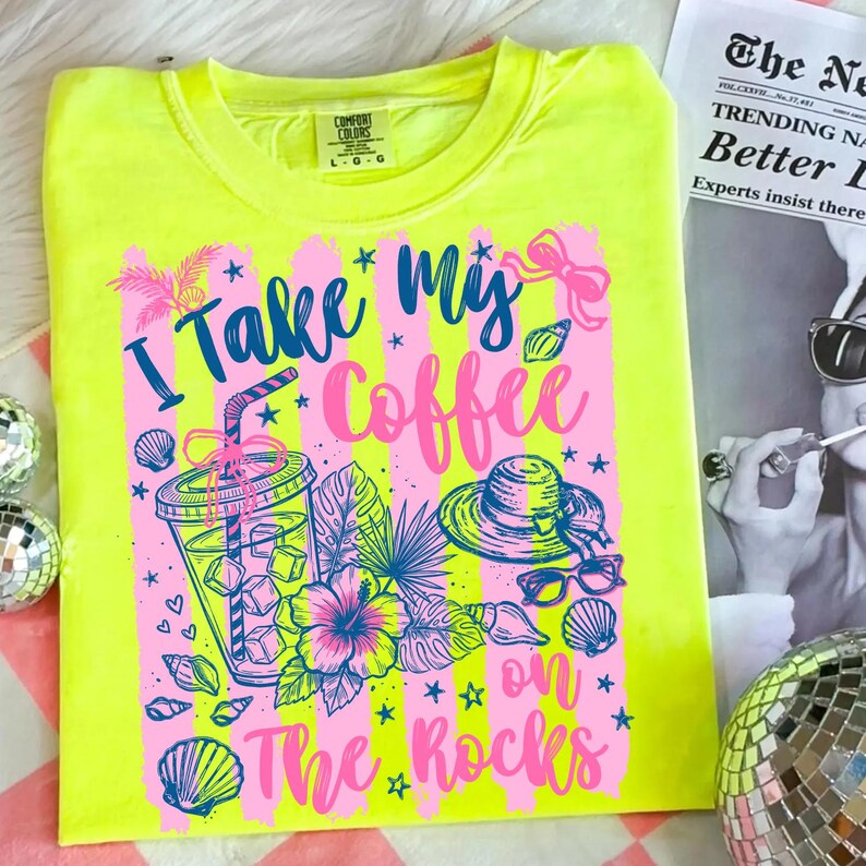 May include: Neon yellow t-shirt with a graphic design that says "I Take My Coffee on The Rocks." The design includes a coffee cup, hat, sunglasses, and tropical flowers in pink and blue.