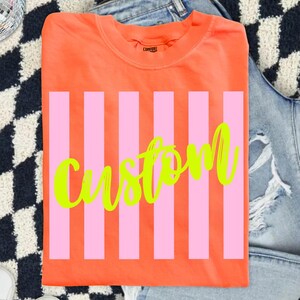 K&ouml;nnte beinhalten: Ein orangefarbenes T-Shirt mit rosa vertikalen Streifen und dem Wort "Custom" in neongr&uuml;ner Schrift. Das T-Shirt ist gefaltet und hat einen Rundhalsausschnitt.