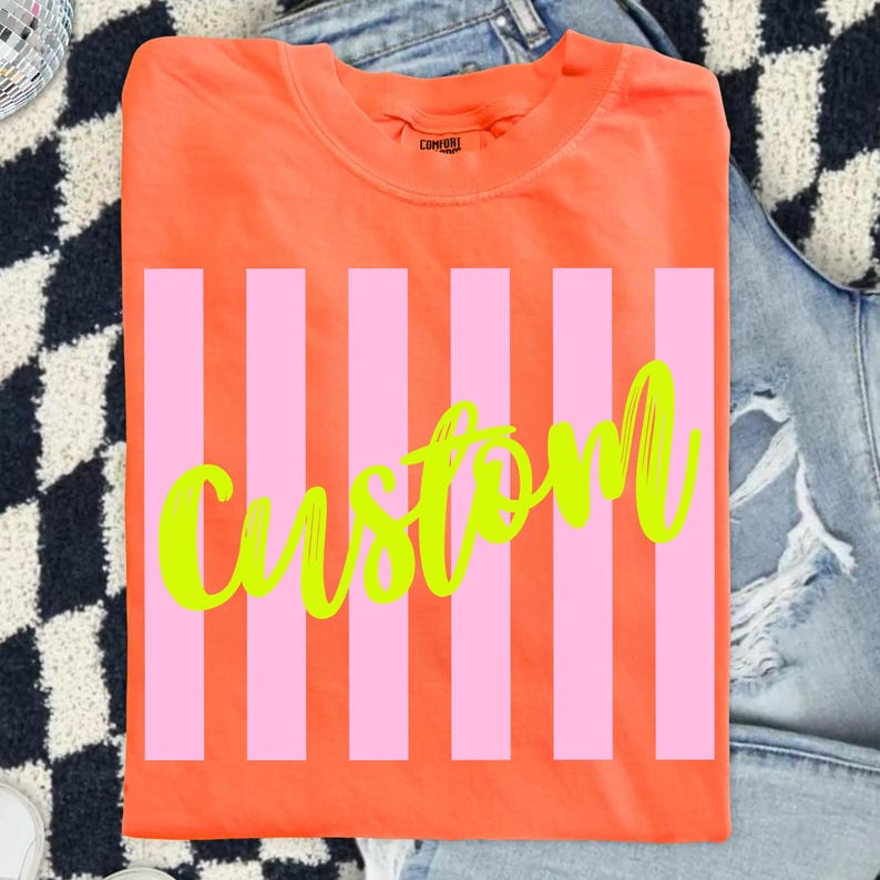 K&ouml;nnte beinhalten: Ein orangefarbenes T-Shirt mit rosa vertikalen Streifen und dem Wort "Custom" in neongr&uuml;ner Schrift. Das T-Shirt ist gefaltet und hat einen Rundhalsausschnitt.