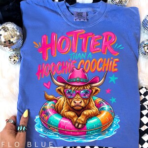 K&ouml;nnte beinhalten: Flo blaues Comfort Colors T-Shirt mit dem Text "Hotter Than a Hoochie Coochie". Die Grafik zeigt eine braune Hochlandkuh mit rosa Cowboyhut und Sonnenbrille, die in einem bunten Schwimmreifen schwimmt. Das Shirt hat eine lockere Passform.