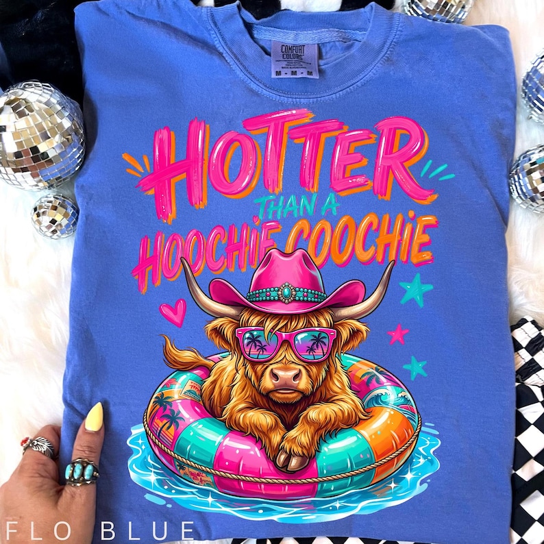 K&ouml;nnte beinhalten: Flo blaues Comfort Colors T-Shirt mit dem Text "Hotter Than a Hoochie Coochie". Die Grafik zeigt eine braune Hochlandkuh mit rosa Cowboyhut und Sonnenbrille, die in einem bunten Schwimmreifen schwimmt. Das Shirt hat eine lockere Passform.