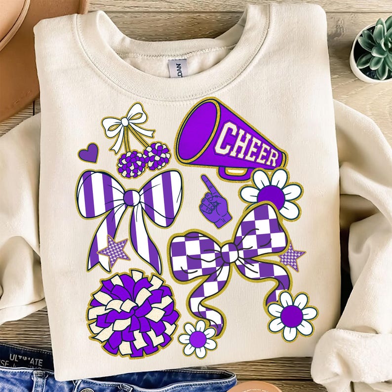 May include: Cream-colored sweatshirt with purple and white cheerleading-themed graphics. Designs include bows, pom-poms, flowers, stars, a megaphone with the word "CHEER", and a pointing hand. The overall design is playful and energetic.