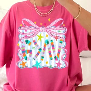 May include: A hot pink t-shirt with a colorful graphic. The graphic features the word "GIV" in rainbow stripes, surrounded by a pink bow, stars, and a blue and white striped background. The shirt is a casual style.
