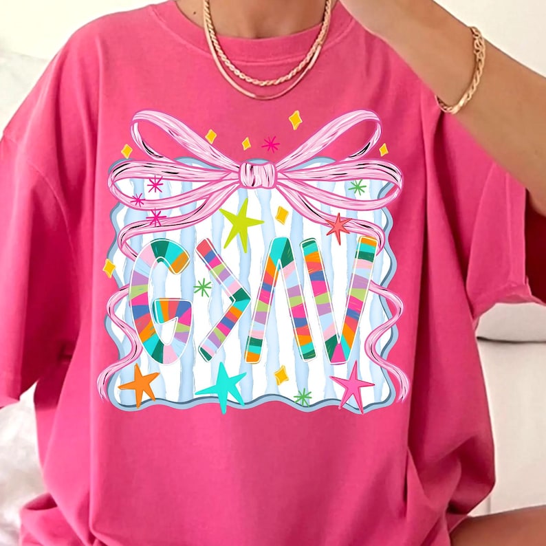 May include: A hot pink t-shirt with a colorful graphic. The graphic features the word "GIV" in rainbow stripes, surrounded by a pink bow, stars, and a blue and white striped background. The shirt is a casual style.