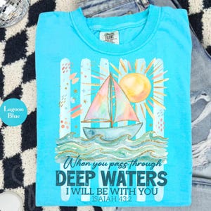 K&ouml;nnte beinhalten: Lagunenblaues T-Shirt mit Segelboot-Design, Regenbogen-Segel, Sonne und Wellen. Der Text auf dem Shirt lautet: "Wenn du durch tiefe Wasser gehst, bin ich bei dir, Jesaja 43,2."