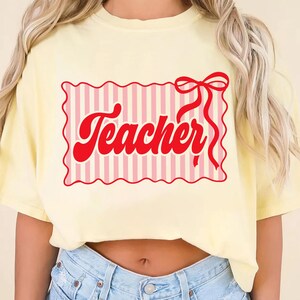 May include: A light yellow t-shirt with a red graphic that says "Teacher". The graphic is in a retro font and is inside a pink and white striped rectangle with a red bow. The shirt is a casual style.