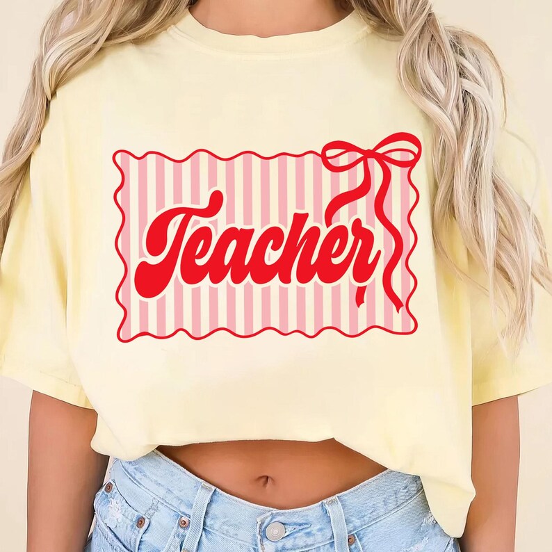 May include: A light yellow t-shirt with a red graphic that says "Teacher". The graphic is in a retro font and is inside a pink and white striped rectangle with a red bow. The shirt is a casual style.
