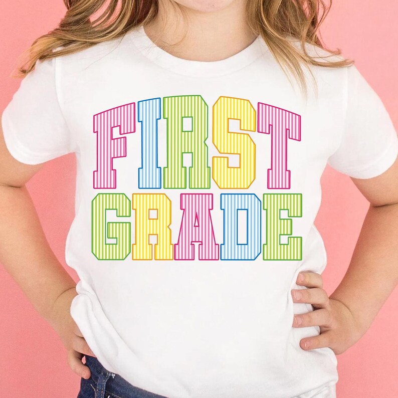 May include: A white short-sleeved t-shirt featuring the text FIRST GRADE printed in a bold, block-style font. Each letter is filled with vertical stripes in alternating bright colors including pink, blue, yellow, and green.