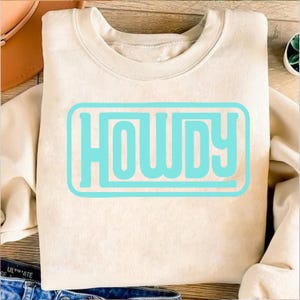May include: A cream-colored sweatshirt with the word "HOWDY" in a light blue rectangular design. The sweatshirt is folded and placed on a wooden surface. The word "HOWDY" is in a bold, sans-serif font.