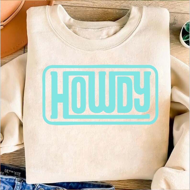 May include: A cream-colored sweatshirt with the word "HOWDY" in a light blue rectangular design. The sweatshirt is folded and placed on a wooden surface. The word "HOWDY" is in a bold, sans-serif font.