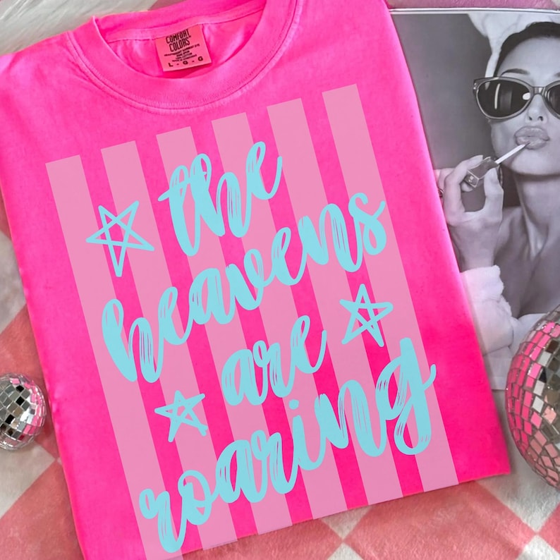 May include: A neon pink t-shirt with vertical light pink stripes and the text "the heavens are roaring" in turquoise script. The shirt has a crew neck and short sleeves. The Comfort Colors brand label is visible.