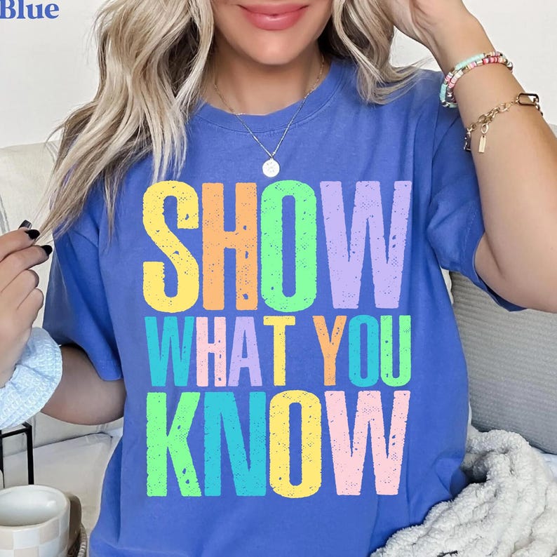May include: A blue t-shirt with the words "SHOW WHAT YOU KNOW" in large, colorful letters. The text is in a variety of colors, including yellow, orange, green, and purple. The shirt is a solid blue color.