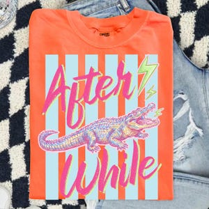 Pode incluir: Camiseta laranja com um design gr&aacute;fico com um jacar&eacute; e as palavras "After While" em rosa e verde. O design inclui listras verticais azuis e raios.
