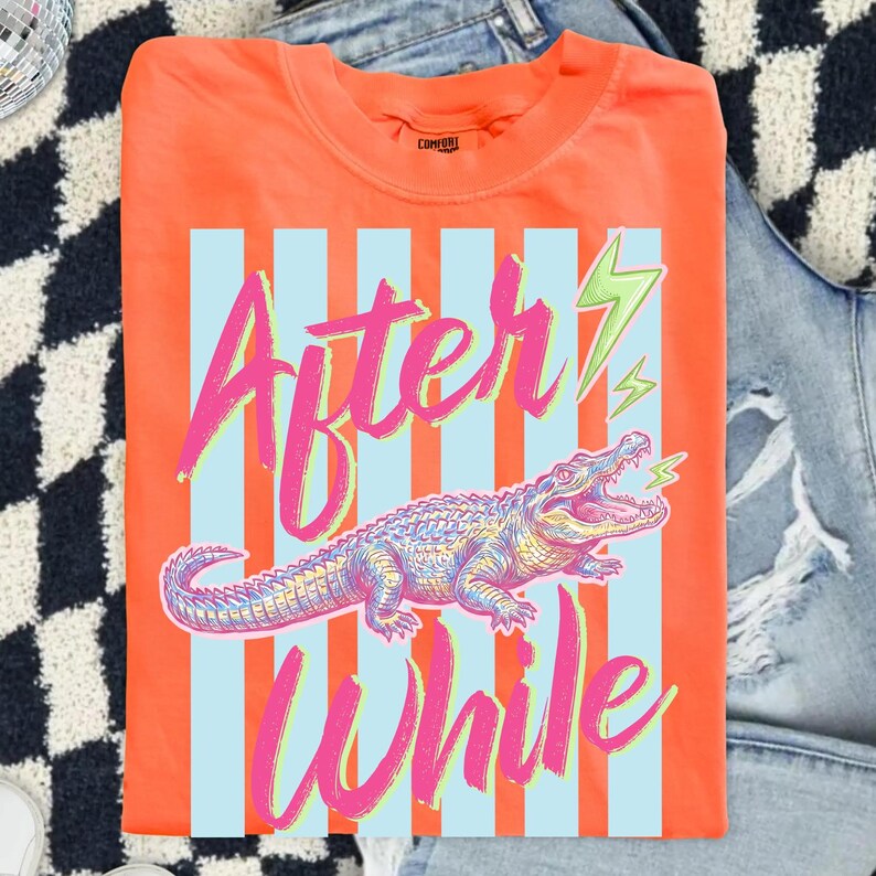 Pode incluir: Camiseta laranja com um design gr&aacute;fico com um jacar&eacute; e as palavras "After While" em rosa e verde. O design inclui listras verticais azuis e raios.