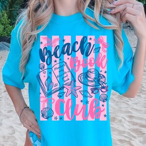 May include: A turquoise t-shirt with a pink and white striped design featuring the words "Beach Book Club." The graphic includes illustrations of a book, seashells, a starfish, sunglasses, and a sun hat. The text "Members only dive into a good book" is also included.