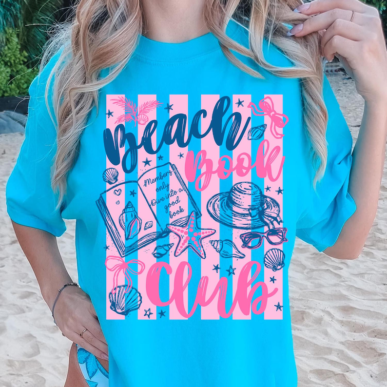May include: A turquoise t-shirt with a pink and white striped design featuring the words "Beach Book Club." The graphic includes illustrations of a book, seashells, a starfish, sunglasses, and a sun hat. The text "Members only dive into a good book" is also included.
