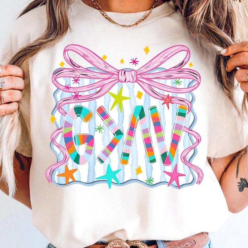 May include: Cream-colored t-shirt featuring a colorful graphic design. The design includes the word "GAV" in vibrant, striped letters, a pink bow, and various stars. The background has blue and white vertical stripes.