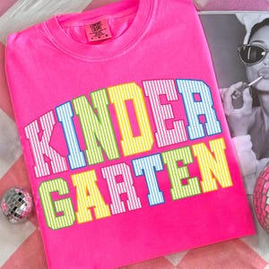 May include: A bright neon pink t-shirt featuring the word KINDERGARTEN printed in large, arched block letters. The letters are filled with a vertical white striped pattern and alternate between light blue, lime green, and yellow colors.