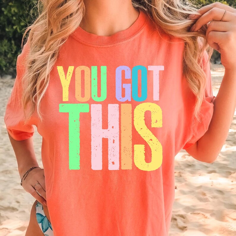 May include: Coral-colored t-shirt with the phrase "YOU GOT THIS" in large, colorful block letters. The text is in a variety of pastel colors, including yellow, green, pink, and blue. The shirt has a crew neck and short sleeves.