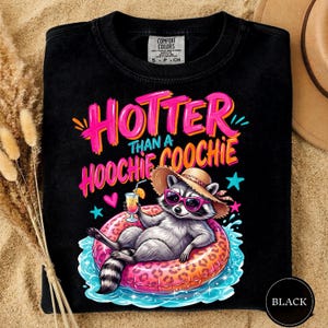 K&ouml;nnte beinhalten: Schwarzes T-Shirt mit dem Schriftzug "HOTTER THAN A HOOCHIE COOCHIE" in Pink und Orange. Ein Waschb&auml;r mit Sonnenbrille und Strohhut entspannt sich auf einem pink-orangenen Schwimmring und h&auml;lt einen Cocktail. Das T-Shirt hat ein Comfort Colors-Etikett.