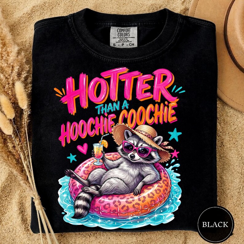 K&ouml;nnte beinhalten: Schwarzes T-Shirt mit dem Schriftzug "HOTTER THAN A HOOCHIE COOCHIE" in Pink und Orange. Ein Waschb&auml;r mit Sonnenbrille und Strohhut entspannt sich auf einem pink-orangenen Schwimmring und h&auml;lt einen Cocktail. Das T-Shirt hat ein Comfort Colors-Etikett.