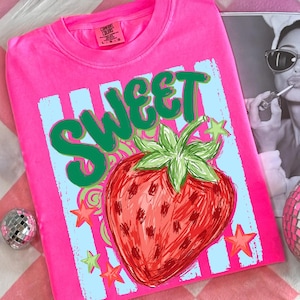 May include: A bright pink t-shirt with a graphic design featuring the word "SWEET" in green, a red strawberry, and stars. The shirt has a crew neck and short sleeves. The brand tag is visible on the upper left.