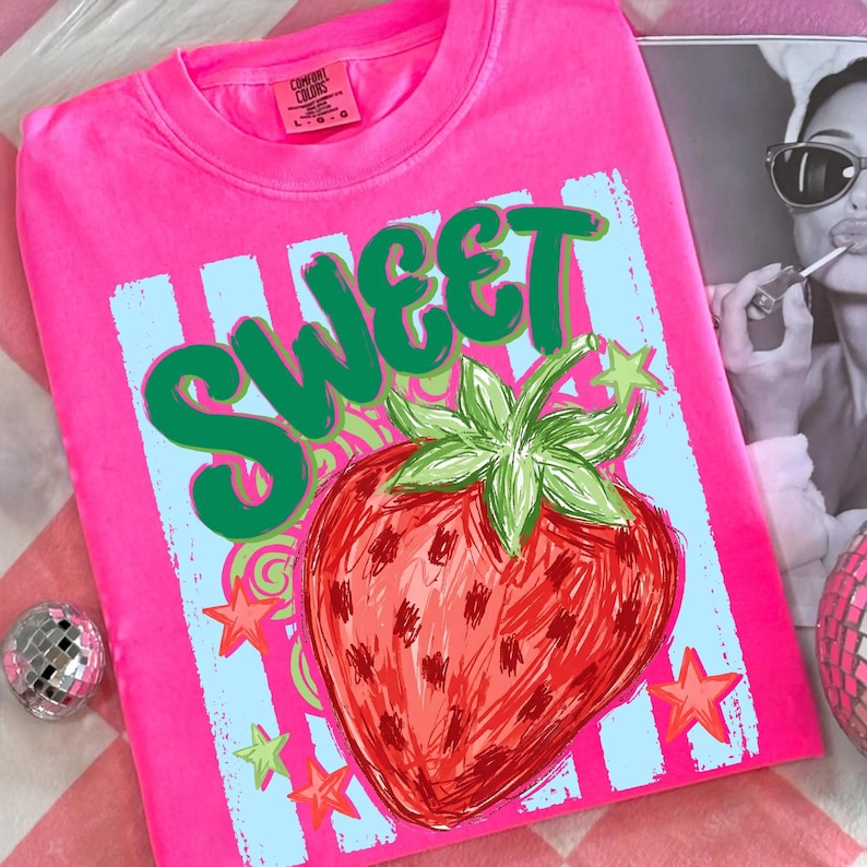 May include: A bright pink t-shirt with a graphic design featuring the word "SWEET" in green, a red strawberry, and stars. The shirt has a crew neck and short sleeves. The brand tag is visible on the upper left.