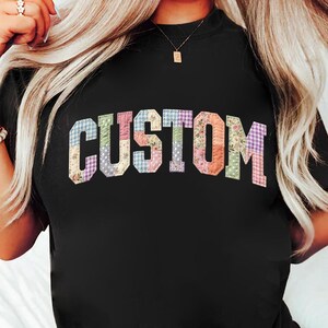 May include: Black t-shirt with the word "CUSTOM" in a patchwork design. The letters are made of various fabrics with floral, gingham, and solid patterns in pastel colors. The shirt has a crew neck.
