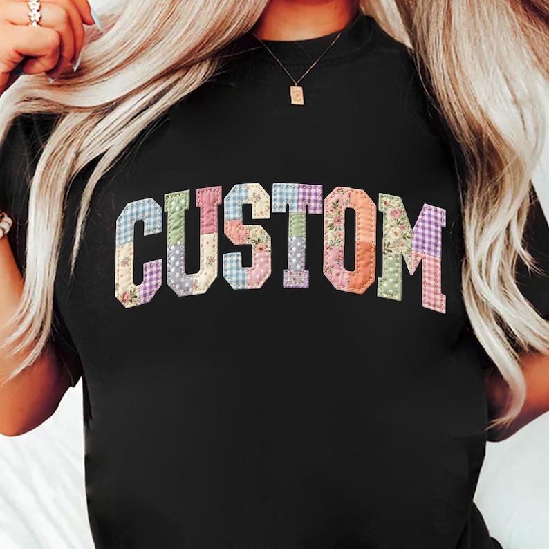 May include: Black t-shirt with the word "CUSTOM" in a patchwork design. The letters are made of various fabrics with floral, gingham, and solid patterns in pastel colors. The shirt has a crew neck.